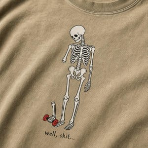 Pode incluir: Uma t-shirt bege com um gráfico de um esqueleto com um braço separado segurando um haltere vermelho. O esqueleto olha para baixo e o texto "well, shit..." está impresso em tinta preta abaixo da figura no tecido da peça de vestuário.