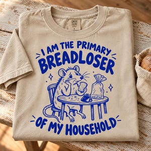 May include: A beige t-shirt featuring a blue graphic of a mouse sitting at a table with a slice of bread and a small purse. The text reads I AM THE PRIMARY BREADLOSER OF MY HOUSEHOLD. The shirt has a Comfort Colors label at the inner neckline.