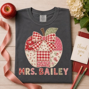 May include: A dark grey t-shirt featuring a large apple-shaped appliqué made of various red, white, and pink floral and gingham fabric patches. A red and white gingham bow sits atop the apple. The text MRS. BAILEY is printed in red and white.