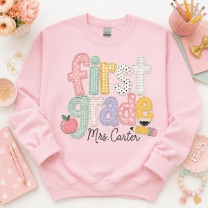 Può includere: Una felpa girocollo rosa chiaro con la scritta First grade in lettere colorate e fantasia in stile ricamato. Il design include una piccola mela rossa e una matita gialla, con il nome Mrs. Carter scritto in corsivo nero sottostante.