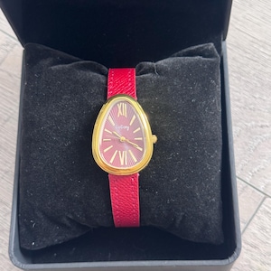Könnte beinhalten: Eine goldfarbene Armbanduhr mit rotem Zifferblatt und passendem roten Lederarmband. Das Zifferblatt hat eine einzigartige ovale Form und verfügt über goldfarbene Zeiger und römische Ziffern. Die Uhr wird in einer schwarzen Box präsentiert.
