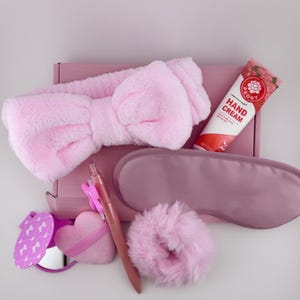 Op de afbeelding: Een roze geschenkset met een pluizige strik-hoofdband, oogmasker, handcrème, pen, spiegel, hartvormige poederdons en scrunchie. De items zijn gerangschikt op een roze doos, wat een samenhangende en vrouwelijke uitstraling creëert.