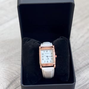 Puede incluir: Un reloj de pulsera rectangular con esfera blanca y marco dorado, exhibido en una caja negra. El reloj tiene una correa de cuero blanca y el texto "WoMaGe" en la esfera. La caja está forrada con terciopelo negro.