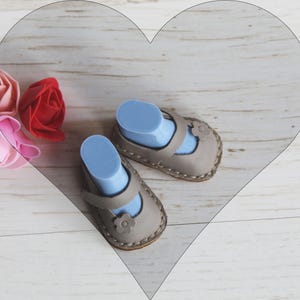 Könnte beinhalten: Ein Paar graue Baby-Lederschuhe mit Riemen und Blumendetail, präsentiert auf blauen Schuhformen. In der Nähe sind rosa und rote Stoffrosen angeordnet, was auf ein Baby- oder Kleinkindthema hindeutet.