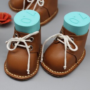 May include: Two brown leather baby booties with white laces. The booties are filled with blue foam blocks that have the number "0" printed on them.