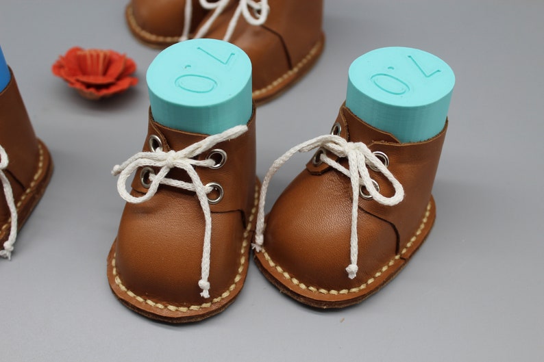 May include: Two brown leather baby booties with white laces. The booties are filled with blue foam blocks that have the number "0" printed on them.