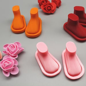May include: Three pairs of miniature doll shoes in orange, red, and pink. The shoes are made of a smooth, rubber-like material and have a rounded toe.