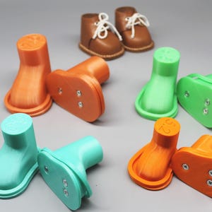 Peut inclure: Diverses pièces de chaussures de poupée imprimées en 3D aux couleurs vives : orange, vert et sarcelle. Deux paires de bottes de poupée en cuir marron avec des lacets blancs sont à l'arrière-plan. Les composants comprennent des bases et des pièces en forme de botte.