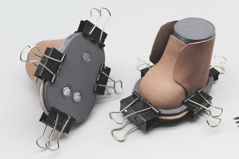 May include: Two brown leather shoe forms are being held together with binder clips. The forms are being used to create a shoe.