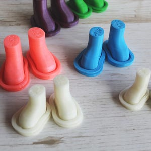 Może przedstawiać: Kolekcja małych, wydrukowanych w 3D plastikowych figurek w kształcie butów w różnych kolorach, w tym neonowy pomarańczowy, jasnoniebieski, złamany biały, fioletowy i zielony. Każda para butów ma gładkie, matowe wykończenie i wyraźną podeszwę.