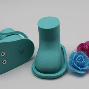 May include: Two turquoise blue 3D printed shoe forms with a base and a raised top. The shoe forms are on a white surface with three artificial flowers in pink and blue.