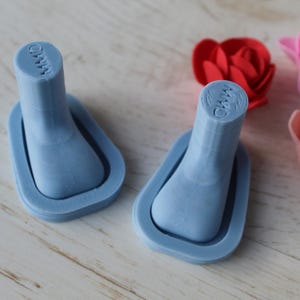 May include: Two light blue 3D-printed objects with a rectangular base and a cylindrical top, each with the letters "MMO" on top. Red, pink, and peach-colored artificial roses are in the background. The objects are on a light wood surface.