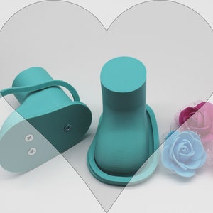 May include: Two teal blue baby shoes with a light blue sole. The shoes have a strap around the ankle and two small screws on the bottom. The shoes are sitting on a white surface with three artificial flowers.