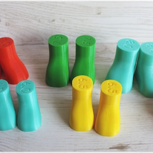May include: A collection of colorful shoe lasts in red, green, yellow, and teal. Each last is labeled with a size, such as 6.0, 6.5, and 7.5. The lasts are arranged on a light-colored wooden surface.