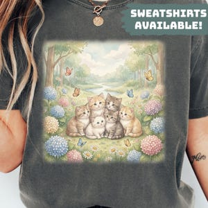 Peut inclure: Sweat-shirt gris foncé avec une illustration à l'aquarelle de sept chatons dans une scène de forêt avec des papillons, des fleurs et une rivière. Le texte "SWEATSHIRTS AVAILABLE!" est imprimé en haut. Le design est doux et fantaisiste.