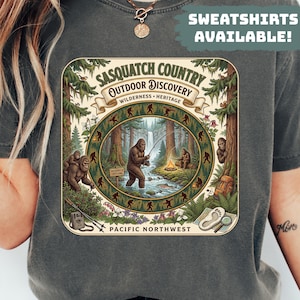 May include: Dark grey t-shirt featuring a detailed illustration of a Bigfoot scene. The design includes text that reads "Sasquatch Country Outdoor Discovery" and "Pacific Northwest". The image depicts Bigfoot fishing, with a campfire and forest setting.