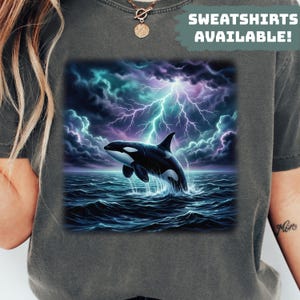 Può includere: Felpa grigio scuro con una grafica di un'orca bianca e nera che emerge dall'oceano sotto un cielo tempestoso con fulmini. Il testo "SWEATSHIRTS AVAILABLE!" è stampato in bianco.