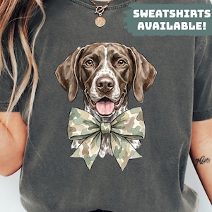 Op de afbeelding: Donkergrijs sweatshirt met een gedetailleerde illustratie van een Duitse Staande Hond met een camouflage vlinderdas. De hond heeft bruine en witte markeringen. Tekst rechtsboven: "SWEATSHIRTS AVAILABLE!"