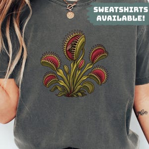 Może przedstawiać: Ciemnoszary t-shirt z graficzną ilustracją muchołówki z zielonymi liśćmi i czerwonymi pułapkami, z których jedna zawiera małego owada. Napis SWEATSHIRTS AVAILABLE! jest wydrukowany białymi literami na zielonym tle.