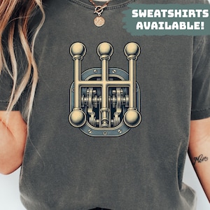 May include: A charcoal gray t-shirt featuring a graphic of a mechanical gear shifter with three vertical levers and rounded knobs in a metallic gold finish. The text SWEATSHIRTS AVAILABLE! is printed in white on a muted green background.