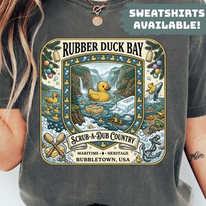 May include: Dark gray t-shirt featuring a colorful graphic with the text "RUBBER DUCK BAY" and "SCRUB-A-DUB COUNTRY". The design includes a yellow rubber duck, waterfalls, and other nautical elements. The text "SWEATSHIRTS AVAILABLE!" is also visible.