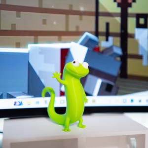 May include: A bright green lizard figurine with a friendly wave. The toy lizard has large, round eyes and is standing on a white shelf. The background includes a computer screen with a pixelated design.
