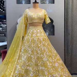 May include: A yellow lehenga, a traditional Indian garment, displayed on a mannequin. The outfit features a short-sleeved top and a long skirt, both heavily embellished with silver and white floral patterns. A matching sheer dupatta is draped over the mannequin's arm.