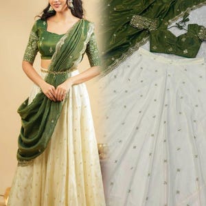 Puede incluir: Un atuendo tradicional indio en verde oliva y crema. El conjunto incluye un top de manga corta, una falda larga y una bufanda drapeada. La falda color crema tiene detalles dorados. El top y la bufanda son de color verde oliva con detalles dorados.