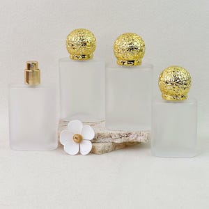Op de afbeelding: Vier rechthoekige parfumflesjes van matglas. Drie flesjes hebben sierlijke, bolvormige gouden doppen met ingewikkelde reliëfpatronen. Eén flesje heeft een eenvoudige gouden verstuiver. Een kleine witte bloemversiering ligt aan de voorzijde.