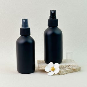 Puede incluir: Dos botellas de spray negro mate con boquillas de spray negras. Una botella tiene una tapa transparente. Las botellas están sobre una superficie de piedra de color claro, con una flor blanca con centro amarillo. Las botellas son cilíndricas.