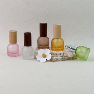 May include: Five small glass bottles with wooden caps in pink, frosted white, brown, and yellow, plus a green bottle with a silver spray nozzle. A white flower ornament sits in the center, arranged with the bottles on a textured stone base.