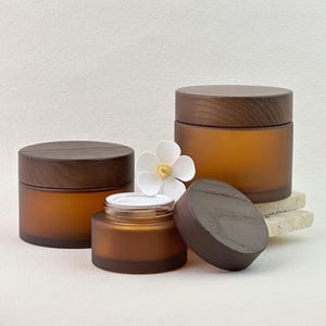 Könnte beinhalten: Drei bernsteinfarbene Glasbehälter mit Holzdeckeln. Ein Behälter ist geöffnet und zeigt einen weißen Innenraum. Eine weiße Blume mit gelbem Zentrum befindet sich zwischen den Behältern. Die Behälter stehen auf einer hellen Oberfläche.