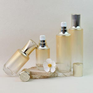 Könnte beinhalten: Eine Auswahl eleganter, klarer Kosmetikflaschen aus Glas mit goldfarbenen Kappen und Akzenten. Die Flaschen variieren in Größe und Form und weisen einen Farbverlauf auf. Eine weiße Blume sorgt für einen dekorativen Touch. Auf neutralem Hintergrund.