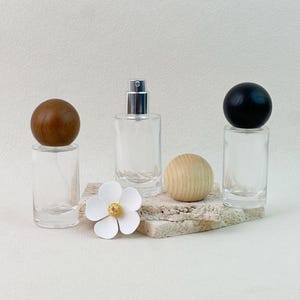 Peut inclure: Flacons de parfum en verre transparent avec des bouchons sphériques en bois et noirs. Une fleur blanche et une sphère en bois sont également présentes. Les flacons sont présentés sur une surface texturée de couleur claire, suggérant une esthétique minimaliste.
