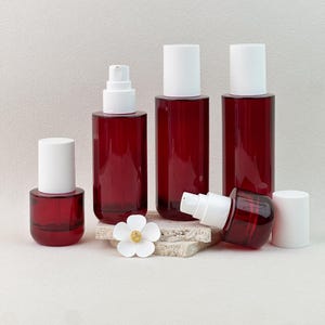 May include: A collection of red glass cosmetic bottles with white caps and pumps. The bottles vary in size and shape, with a small, round bottle, several cylindrical bottles, and one bottle lying on its side. A white flower is placed in front of the bottles.