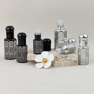 May include: A collection of six hexagonal glass roll-on bottles featuring intricate silver filigree patterns. Three bottles are black with matching caps, while three are silver with metallic caps. One silver bottle lies horizontally.