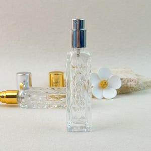 Single silver cap clear glass perfume bottle, front view showing diamond-cut texture, blurred background with second gold cap bottle.
