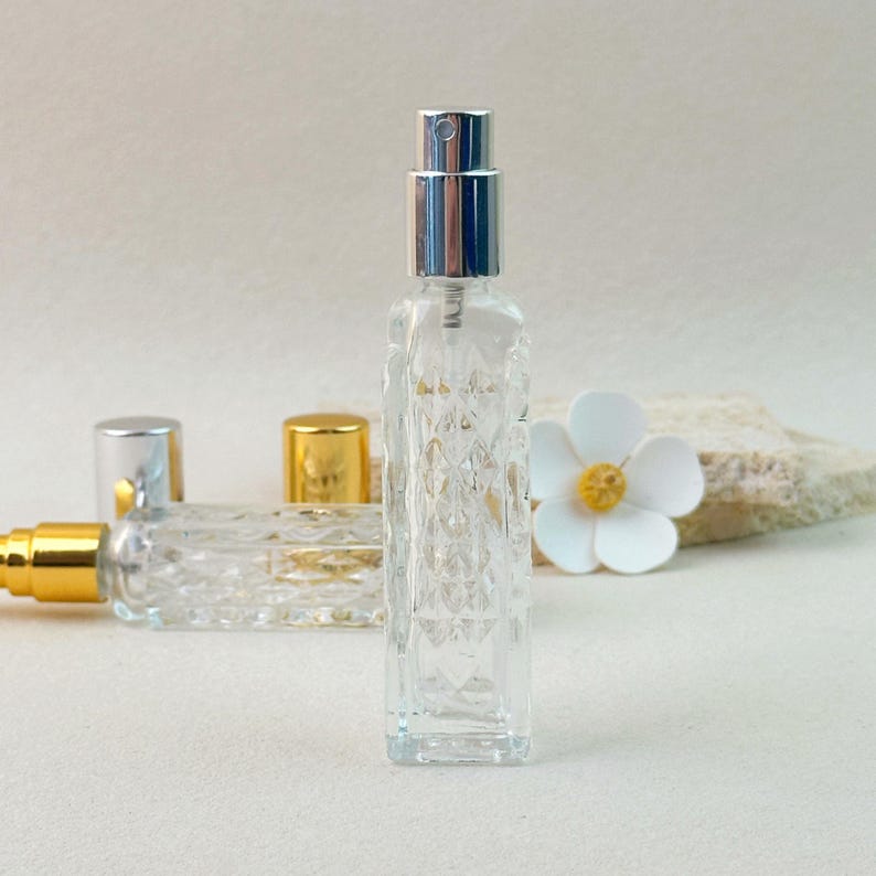 Single silver cap clear glass perfume bottle, front view showing diamond-cut texture, blurred background with second gold cap bottle.