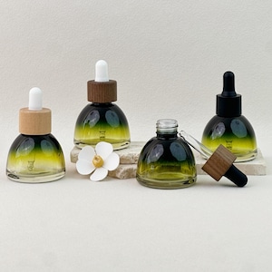 May include: Four glass dropper bottles with a gradient green and yellow color scheme. The bottles have wooden and black caps. One bottle is open, with the dropper separate. A white flower is in the foreground.