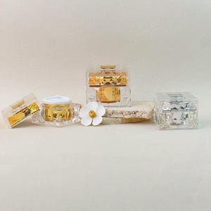 Pode incluir: Uma variedade de pequenos frascos transparentes e facetados com detalhes dourados e prateados. Um frasco tem uma tampa branca. Uma flor branca com centro amarelo também está presente. Os frascos estão dispostos sobre um fundo neutro.