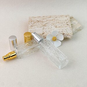 Two perfume atomizer bottles lying flat, silver and gold pumps, with removed caps and flower decor on stone background.