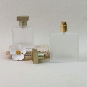 Op de afbeelding: Twee vierkante parfumflesjes van matglas met goudkleurige doppen. Eén flesje is afgesloten, terwijl de andere open is en een gouden verstuiver laat zien. Een witte bloem met vijf blaadjes en een geel hartje ligt naast de flesjes.