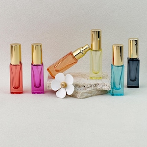 Könnte beinhalten: Eine Sammlung von sechs rechteckigen Glasparfümflaschen mit goldenen Verschlüssen in leuchtenden Farben wie Rot, Pink, Orange, Gelb, Blau und Schwarz. Ein weißes blumenförmiges Ornament befindet sich vor der orangefarbenen Flasche.