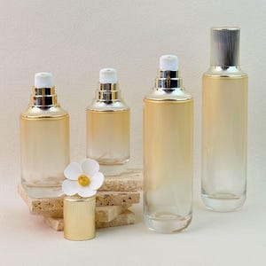 May include: Four glass cosmetic bottles with a gradient peach to clear finish and gold pump dispensers. One bottle features a white flower cap topper. The bottles vary in height and width, showcasing a sleek design for skincare or beauty products.