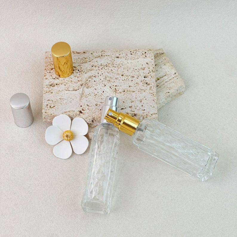Two clear glass perfume bottles with silver and gold caps, tilted view showing pump mechanism on stone background.