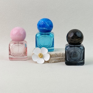 30ml pink, blue, and gray colored glass perfume bottles with marbled caps, minimalist display with flower decor on neutral background.