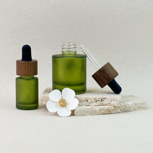 Puede incluir: Dos frascos cuentagotas de vidrio verde oliva esmerilado con cuellos de madera oscura y bulbos de goma negros. Un frasco está cerrado, mientras que el otro está abierto con su pipeta de vidrio retirada. Una flor blanca con centro dorado.