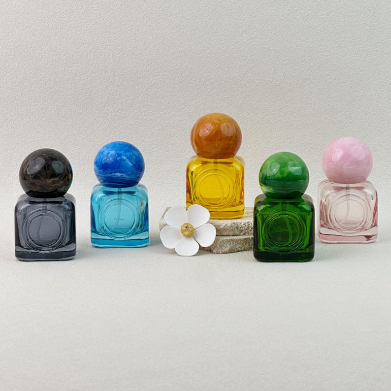 30ml colored glass perfume bottles in pink, yellow, blue, green, gray with matching marbled caps, arranged with flower decor on stone background.