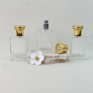 Op de afbeelding: Drie doorzichtige glazen parfumflesjes met een vierkante basis en goudkleurige doppen. Twee flesjes zijn afgesloten, terwijl één gouden dop los op een getextureerd stenen blok ligt. Een wit bloemvormig decoratief accent ligt bij de flesjes.