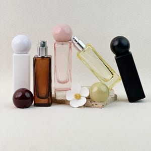 May include: An assortment of perfume bottles in various colors and shapes. The bottles include a white, brown, and pink rectangular bottle, and a yellow and black rectangular bottle. A white flower and a green sphere are also present.